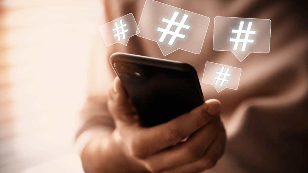 Close-up of a person holding a black cell phone. Hashtags float out of the top of the phone like bubbles. The hashtags say "#️⃣" and other unreadable text.