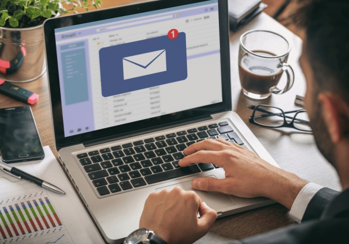 A person working on a laptop displaying an email notification icon. The desk has a coffee mug, smartphone, pen, and papers with graphs. A small plant is visible in the corner of the image.