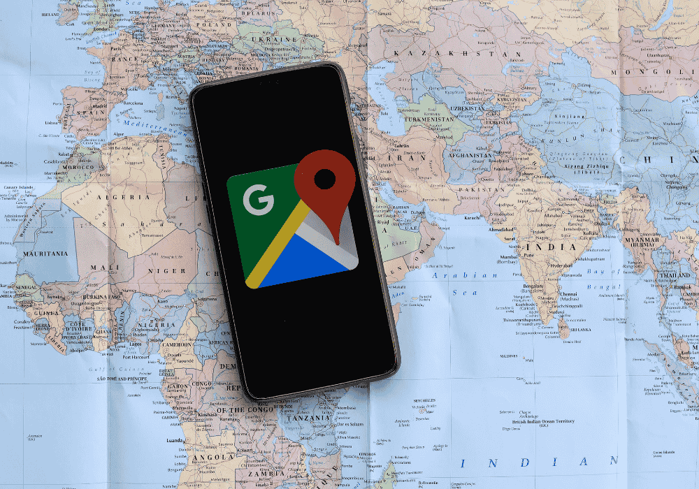 A smartphone screen displaying the Google Maps app icon is placed on top of a world map. The map shows various countries and continents, while the smartphone highlights the Google Maps app, symbolizing navigation and location services.